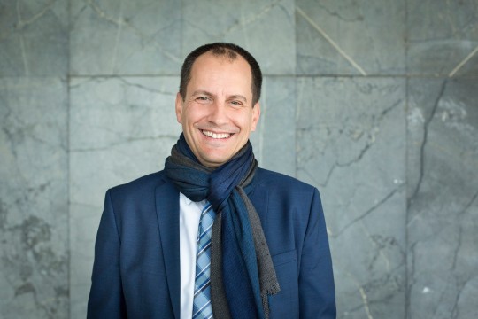 Lächelnder Mann mit Schal, der vor einer Steinwand steht. 