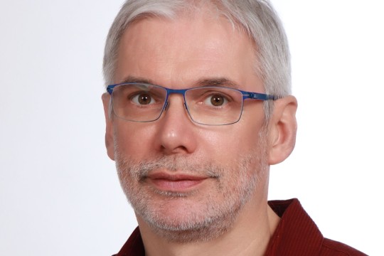 Lächelnder Mann mit Brille vor einem grauen Hintergrund. 