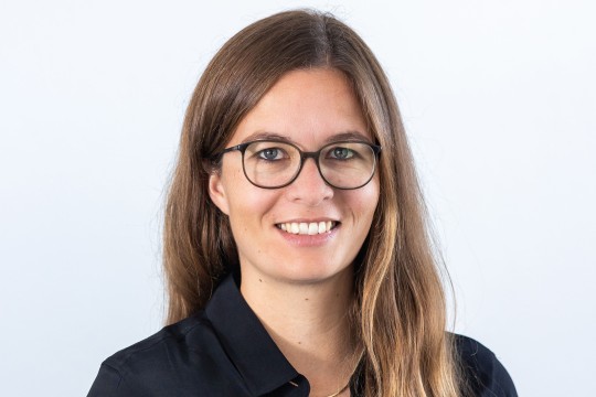 Lächelnde Frau mit Brille vor einem grauen Hintergrund. 