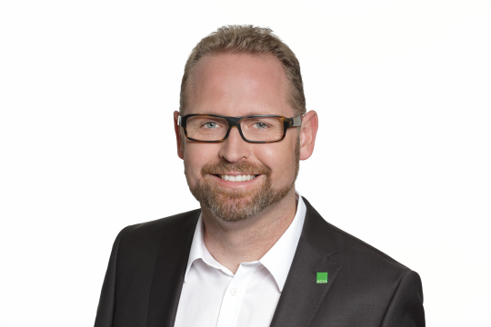 Lächelnder Mann mit dunklem Sakko vor einem hellen Hintergrund.