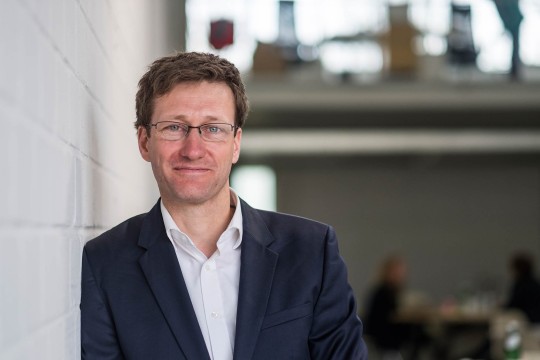 Mann mit einem dunklen Sakko vor einem hellen Hintergrund. 